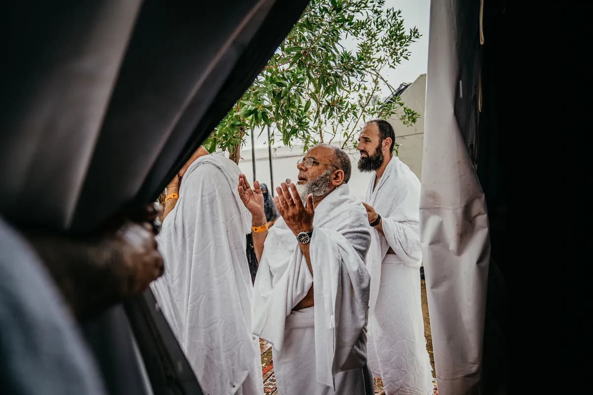 A pilgrim in ihram raises hands in prayer near the Kaaba, surrounded by fellow pilgrims